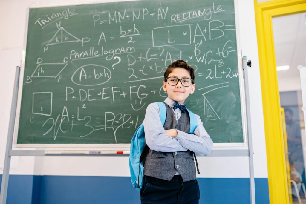 A young student stands confidently with arms crossed in front of a chalkboard filled with math equations.