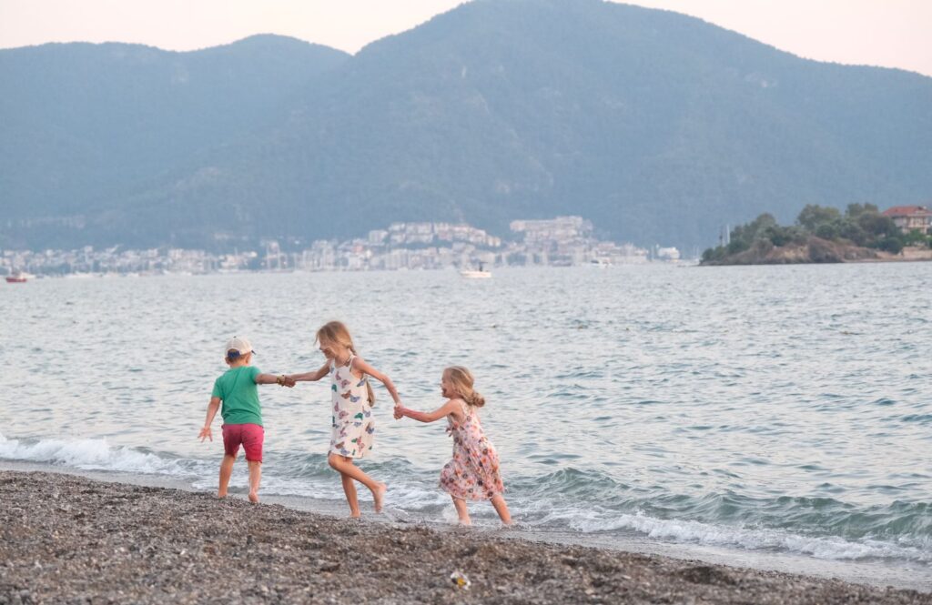 Three children run along the beach in Fethiye against a scenic hillside backdrop.